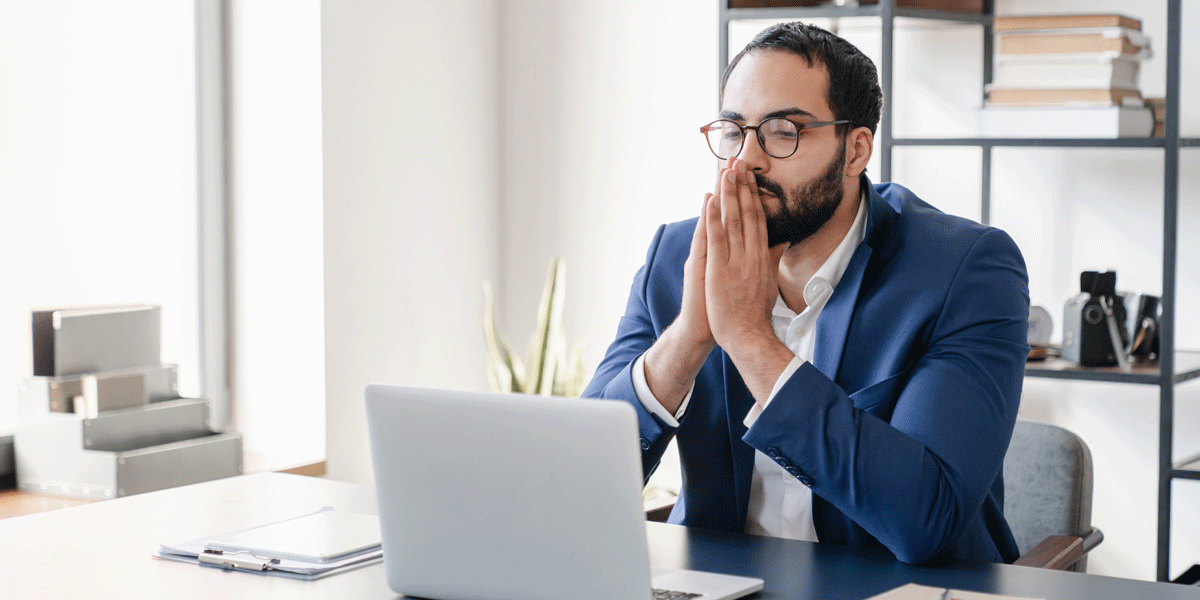Stressed man in business suit sitting at his desk with his hands crossed in a prayer motion in front of his face.